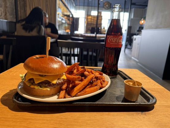 Chilli-Cheeseburger mit Süßkartoffelpommes auf einem Tablett, eine Glasflasche Coca-Cola und ein kleiner Behälter mit Soße, mit Menschen im Hintergrund eines Restaurants.
